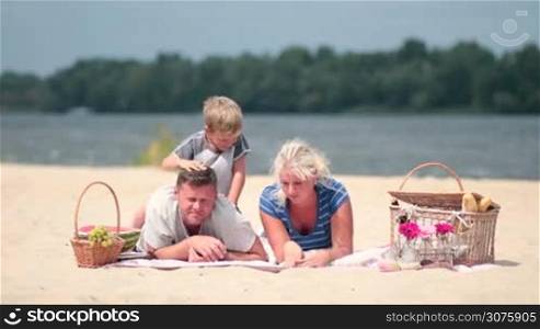 Happy family with boy relaxing on the beach and picnicking during weekend. Handsome dad in casual clothes lying on colorful blanket on the sand and his cute little son lying on him and hugging his neck. Son is a pickaback