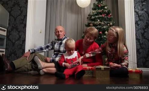 Happy family with children sitting on the floor in living room decorated with Christmas tree ,candles and gift boxes during winter holidays celebration. Granny and toddler grandson in Christmas clothes playing with new toy car.