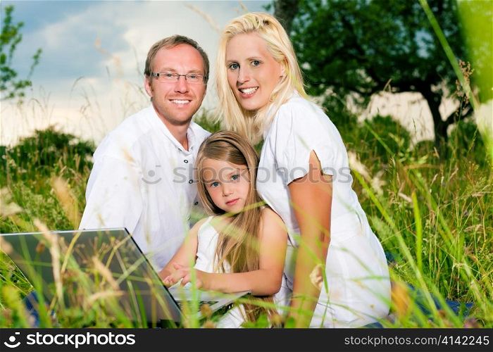 Happy family with daughter girl sitting in a meadow in summer with laptop