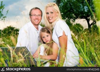 Happy family with daughter girl sitting in a meadow in summer with laptop