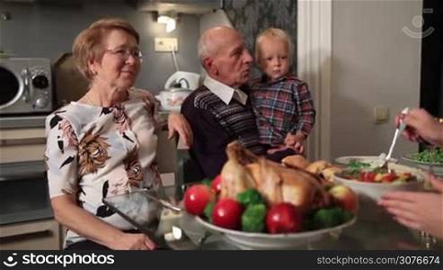 Happy family with grandparents sitting at festive table on Thanksgiving holiday at home. Cute granddaugther in angel dress serving peas to her grandfather and gently kissing him and her little toddler brother while granny talking with adult daugther.