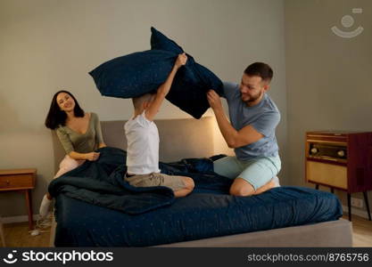 Happy family with small preschooler son have fun enjoy funny activity together. Overjoyed young father and mother with cheerful child engaged in pillow fight on bed. Happy young family in pillow fight at home