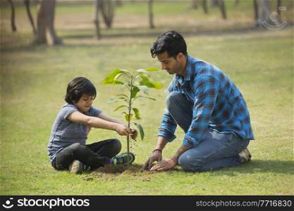 Happy father and son planting a small plant in garden and putting soil around it.