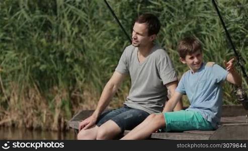 Happy father and son sitting on wooden pier chatting and fishing together on the lake during summer day. Teenage boy and young dad with fish-rods relaxing near the pond while fishing. Slow motion.