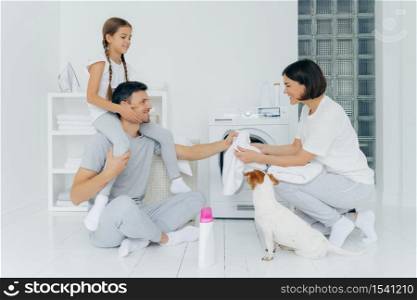 Happy father gives piggy back to daughter, helps wife to load washing machine, their pet poses near, bottle of washing powder in white floor. Friendly family in laundry room, wash linen, do housework