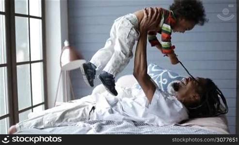 Happy father laying on his back on the bed and raising his cute mixed race child up in the air while his sweet little toddler boy holding daddy&acute;s dreadlocks. Playful handsome african man playing with son in the bedroom at home. Side view.
