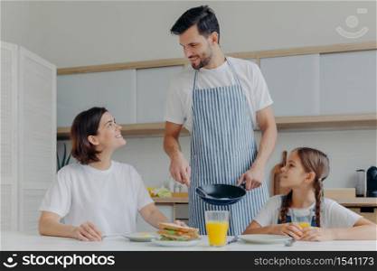 Happy father, mother and daughter gather together at kitchen, eat delicious breakfast, dad prepared fried eggs, being in good mood, ready to start new day. Lovely family enjoy tasty meal at home