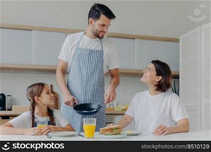 Happy father, mother and daughter gather together at kitchen, eat delicious breakfast, dad prepared fried eggs, being in good mood, ready to start new day. Lovely family enjoy tasty meal at home
