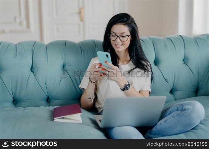 Happy female blogger chats with friends in social netwoks, works on laptop computer, poses in coworking space, sits on sofa in lotus pose, connected to wireless internet. Technology, freelancing