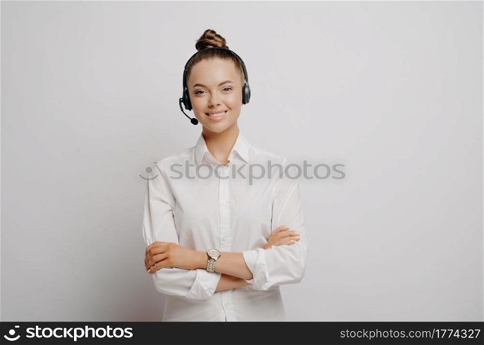 Happy female call center employee in formal wear in black headset feeling happy and assured after learning about promotion, looking at camera with smile while standing alone in front of grey wall. Female call center employee in headset with crossed arms