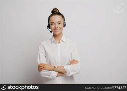 Happy female call center employee in formal wear in black headset feeling happy and assured after learning about promotion, looking at camera with smile while standing alone in front of grey wall. Female call center employee in headset with crossed arms