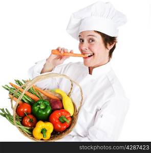 Happy female chef holding a basket of fresh, organic, locally grown produce and eating a raw carrot. Isolated on white background.
