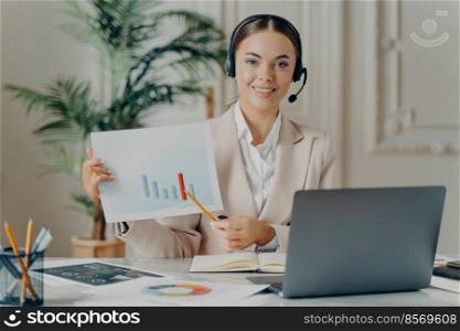 Happy female economist in headset pointing at financial report with rising stats during online meeting on laptop, business woman preparing startup presentation while sitting at her cozy workplace. Business woman in headset with financial document or business plan