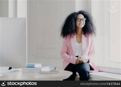 Happy female entrepreneur has bushy Afro hair, wears rosy jacket and black trousers, holds cup of drink, poses at windowsill in spacious cabinet with table, modern computer, enjoys aromatic coffee