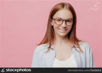 Happy female entrepreneur has straight hair, wears spectacles, smiles positively, enjoys good sales and profits, dressed in stylish shirt, models over pink background with copy space for your text