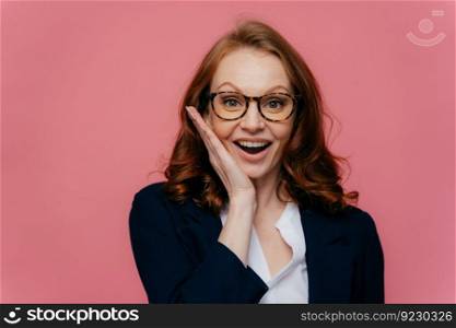 Happy female entreprenuer touches cheek gently, smiles broadly, wears transparent glasses, has ginger hair, dressed in formal apparel, satisfied to achieve success, isolated over pink background