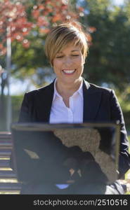 Happy female freelancer keyboards on laptop computer has new ideas for work wearing formal clothes sits on wooden bench outdoor with notebook