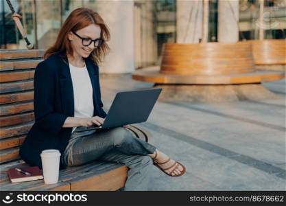 Happy female freelancer keyboards on laptop computer has new ideas for work wears eyeglasses formal clothes sits on wooden bench outdoor with takeaway coffee notebook pen around. Warm sunny day. Happy female freelancer keyboards on laptop computer sits on wooden bench