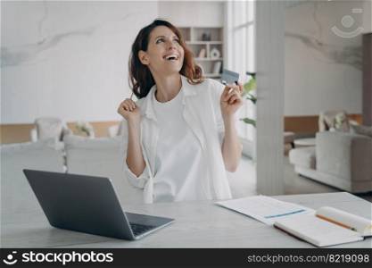 Happy female holding bank credit card got cashback bonus using online banking service at laptop. Hispanic woman excited with easy secure cashless payment. E-banking, e commerce concept.. Happy female holding bank credit card got cashback bonus using online banking service at laptop