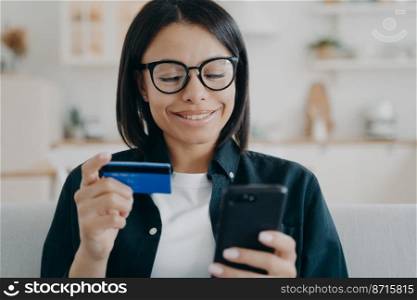 Happy female holding credit card and smartphone, uses online banking services, mobile bank app at home. Young woman making easy secure payment, money transfer, shopping on internet. E-banking.. Happy female holding credit card, smartphone, uses online banking services, mobile bank app at home