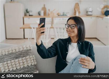 Happy female holding smartphone takes selfie photo or chatting by video call, sitting on sofa at home. Smiling modern young woman wearing glasses communicates online, using mobile phone app.. Happy female holding smartphone takes selfie photo or chatting by video call sitting on sofa at home
