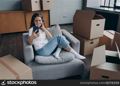 Happy female homeowner tenant holding smartphone chatting with relatives or friends by video call, discuss furniture purchases, relocation, sitting among cardboard boxes with personal belongings.. Happy female homeowner tenant chatting by video call discussing relocation to new home on moving day