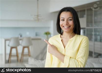 Happy female housewife pointing aside by finger, recommends service, online store offer, standing at home. Smiling pleased woman homeowner sharing shopping discounts, looking at camera indoors.. Happy female housewife pointing aside, recommends service, online store offer, standing at home