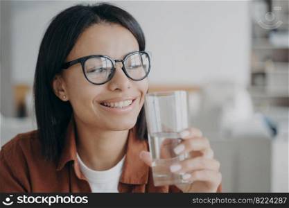 Happy female in glasses holding glass of pure mineral water, looking aside. Smiling woman enjoying clear purified filtered aqua, starts the day with good habit for beauty. Wellness, diet concept.. Happy female in glasses holding glass of pure mineral water. Healthy lifestyle habit, wellness, diet