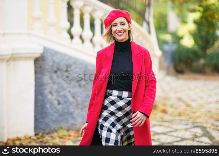Happy female in red outfit and beret looking at camera while strolling on path near fence on street with trees in city. Smiling woman in stylish outfit walking on street