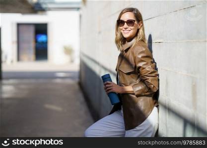 Happy female in stylish clothes with sunglasses and thermos smiling and looking at camera while leaning on wall on sunny day on street. Cheerful stylish woman under sunlight
