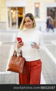Happy female in stylish outfit and handbag browsing modern cellphone while strolling with cup of drink on street. Positive woman with cup of takeaway drink using smartphone