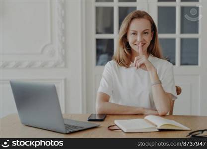 Happy female keep finger at lips asking be quiet and keep secret private information. Smiling young woman makes silence gesture, sitting at office desk with laptop in the workplace.. Smiling woman makes silence gesture, asking to keep secret, sitting at office desk with laptop