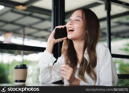 Happy female lifestyle sitting in cafe near window she cellphone on mobile phone, Asian young business woman freelance holding smartphone and coffee paper cup at coffee shop