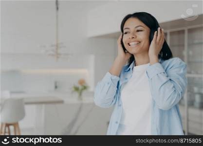 Happy female listening to music or podcast in modern wireless headphones at home. Joyful smiling young woman listens to favorite song, audio book in headset, enjoying perfect sound quality indoors.. Happy female listening to music or podcast in modern wireless headphones, enjoying sound at home