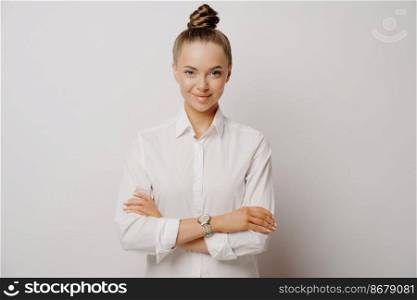 Happy female office employee in formal white shirt expressing confidence, standing with arms crossed while thinking about work day, isolated on light wall. Confident business woman posing in studio. Confident female manager in white shirt with crossed arms