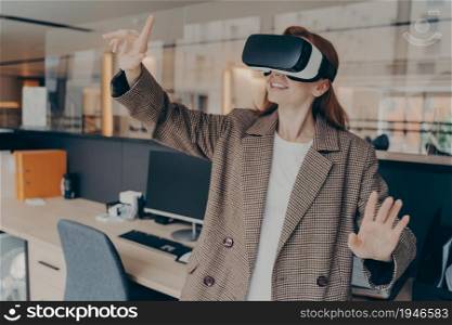 Happy female office worker enjoying virtual reality game on her break, playing using portable VR glasses, moving objects in digital world with her hands, standing in front of her workplace in headset. Happy female office worker in VR headset enjoying virtual reality game on her break