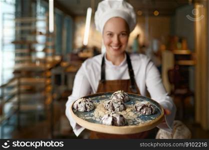 Happy female pastry chef presenting freshly baked cookies standing at bakery kitchen. Happy confectioner showing delicious freshly made sweet brownie on tray. Pastry chef presenting freshly baked cookies at bakery kitchen