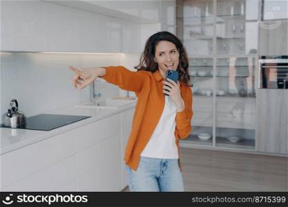 Happy female singing using mobile phone as microphone, having fun, dancing in kitchen. Energetic hispanic woman listening to music, enjoying musical playlist, practicing vocal at home.. Energetic female singing using mobile phone as microphone, having fun, practicing vocal at home