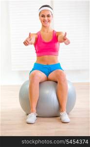 Happy female sitting on fitness ball and showing thumbs up&#xA;