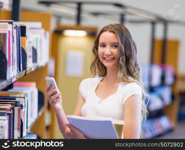 Happy female student holding books at the library. Happy young female student holding books and her mobile phone at the library