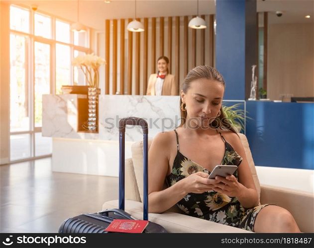 Happy female traveler sitting at hotel lobby with luggage and using smartphone while waiting for hotel check in