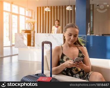 Happy female traveler sitting at hotel lobby with luggage and using smartphone while waiting for hotel check in