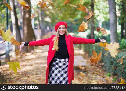 Happy female wearing red beret and coat tossing yellow dry leaves on autumn street and looking at camera. Cheerful woman throwing maple leaves in air