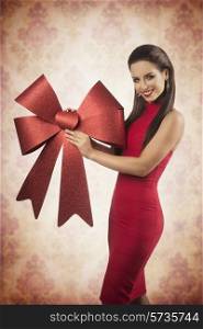 happy female with long brown hair and sexy red dress taking big christmas bow in the hand and smiling