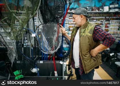 Happy fisherman choosing net in fishing shop, hooks and baubles on background. Equipment and tools for fish catching and hunting, accessory choice on showcase in store, bait assortment. Happy fisherman choosing net in fishing shop