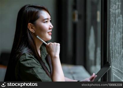 happy freelance asian people business female thinking on something casual working taking note on digital tablet with electronic pen wearing brownish green dress stylish hipster Summer in coffee shop