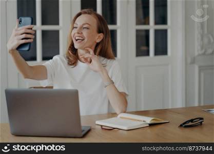 Happy freelance worker girl blogger making phone video call, chatting online, using mobile app on smartphone at desk. Friendly smiling woman takes selfie photo, sitting at laptop in the workplace.. Happy girl blogger makes phone video call, chatting online, takes selfie photo, sitting at laptop