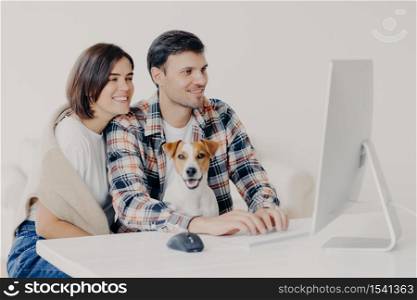 Happy friendly couple sit together with dog, make booking of hotel for future trip, keyboard and look at computer, chat online, pose at white desktop, connected to free wifi, browse internet together