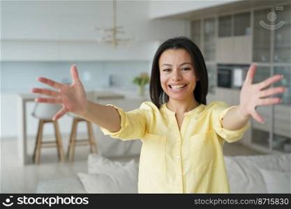 Happy friendly woman in casual clothes stretches arms wide open ready for hug, smiling female looking at camera stretching hands to embrace, showing love, meeting guests, giving warm welcome at home.. Smiling woman stretches arms to hug, looking at camera, meeting guests, giving warm welcome at home