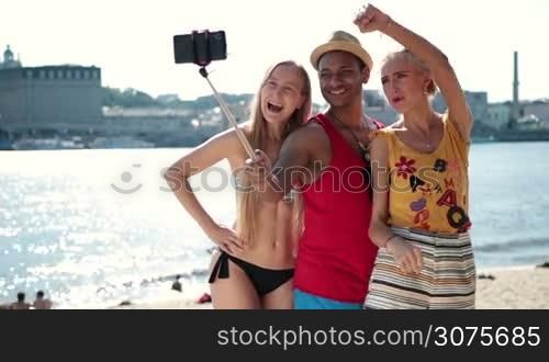 Happy friends making selfie photo on a selfie stick on the beach during summer vacation. Group of smiling friends posing and making selfie on smartphone